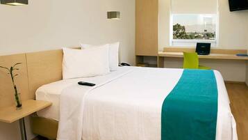 A hotel room with a bed, bedside table, lamp, and a desk with a chair.