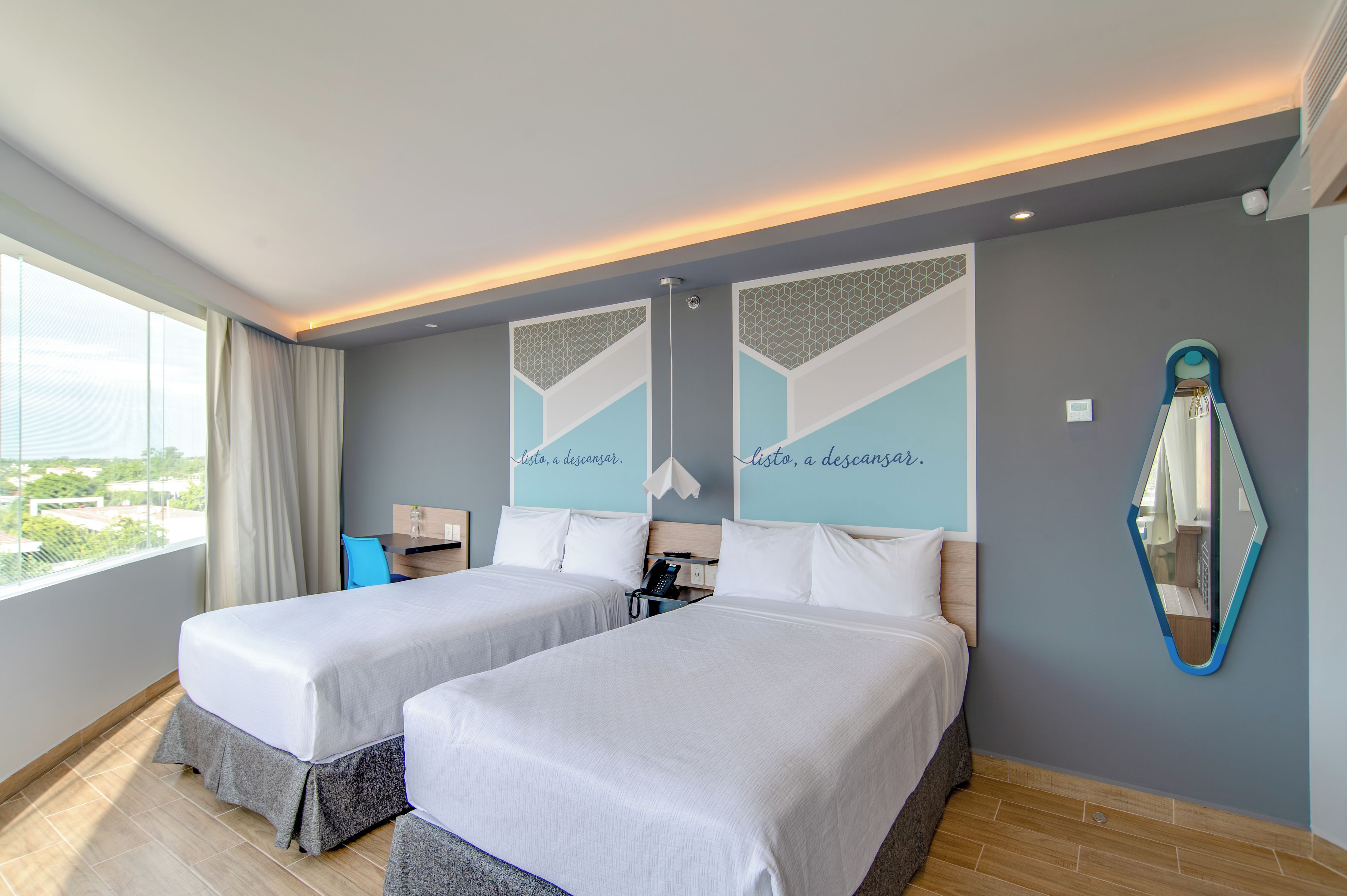 A hotel room with two beds, a desk, a chair, a mirror, and a window with a view.