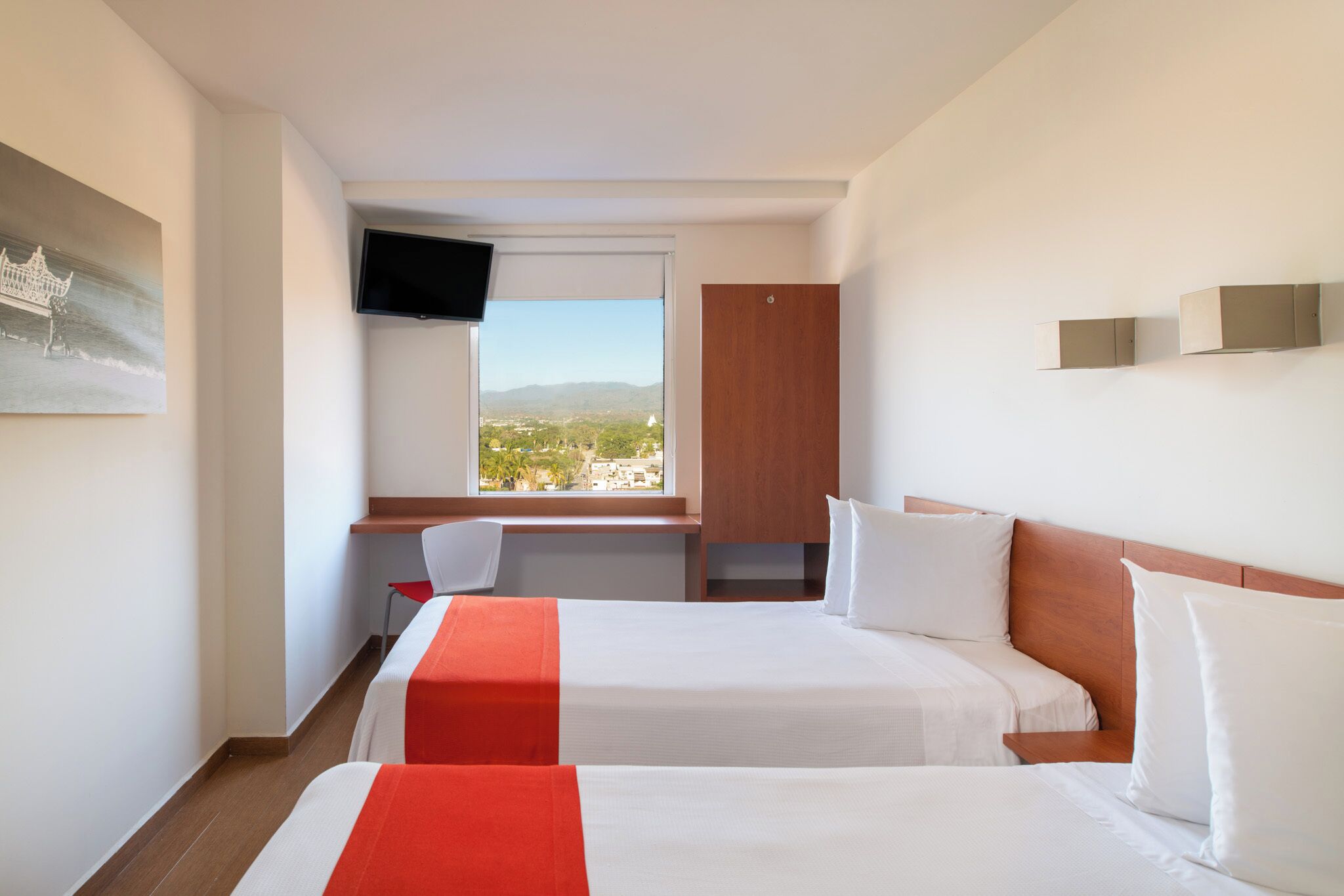 A hotel room with two beds, a desk, a chair, a TV, and a window with a view of the city.