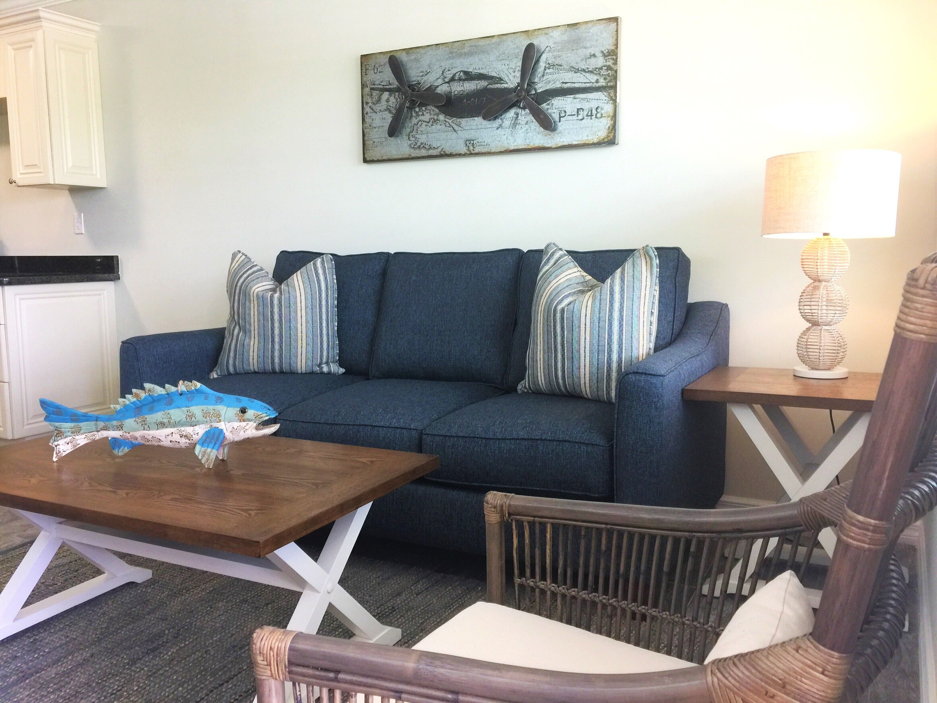 A living room with a blue sofa, striped pillows, a wooden coffee table with a fish model, a wicker chair, a lamp, and a map artwork on the wall.