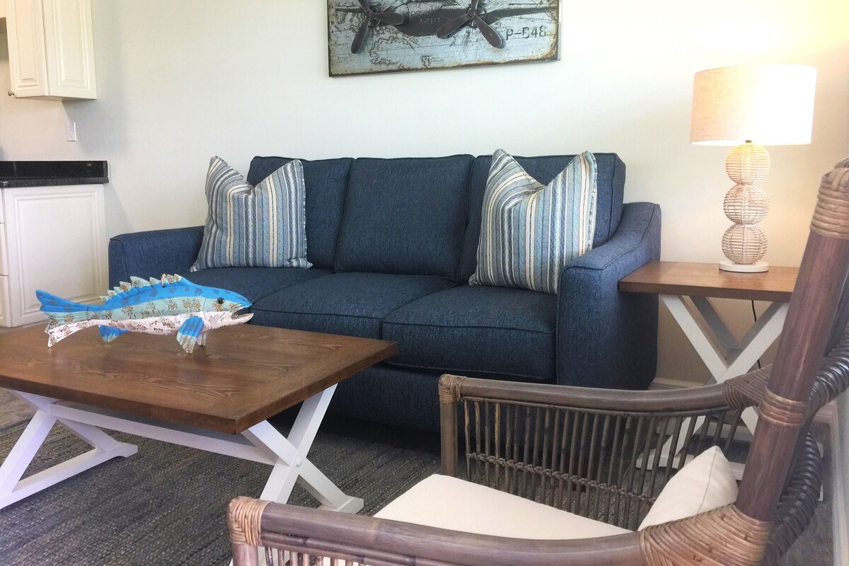 A living room with a blue sofa, striped pillows, a wooden coffee table with a fish model, a wicker chair, a lamp, and a map artwork on the wall.