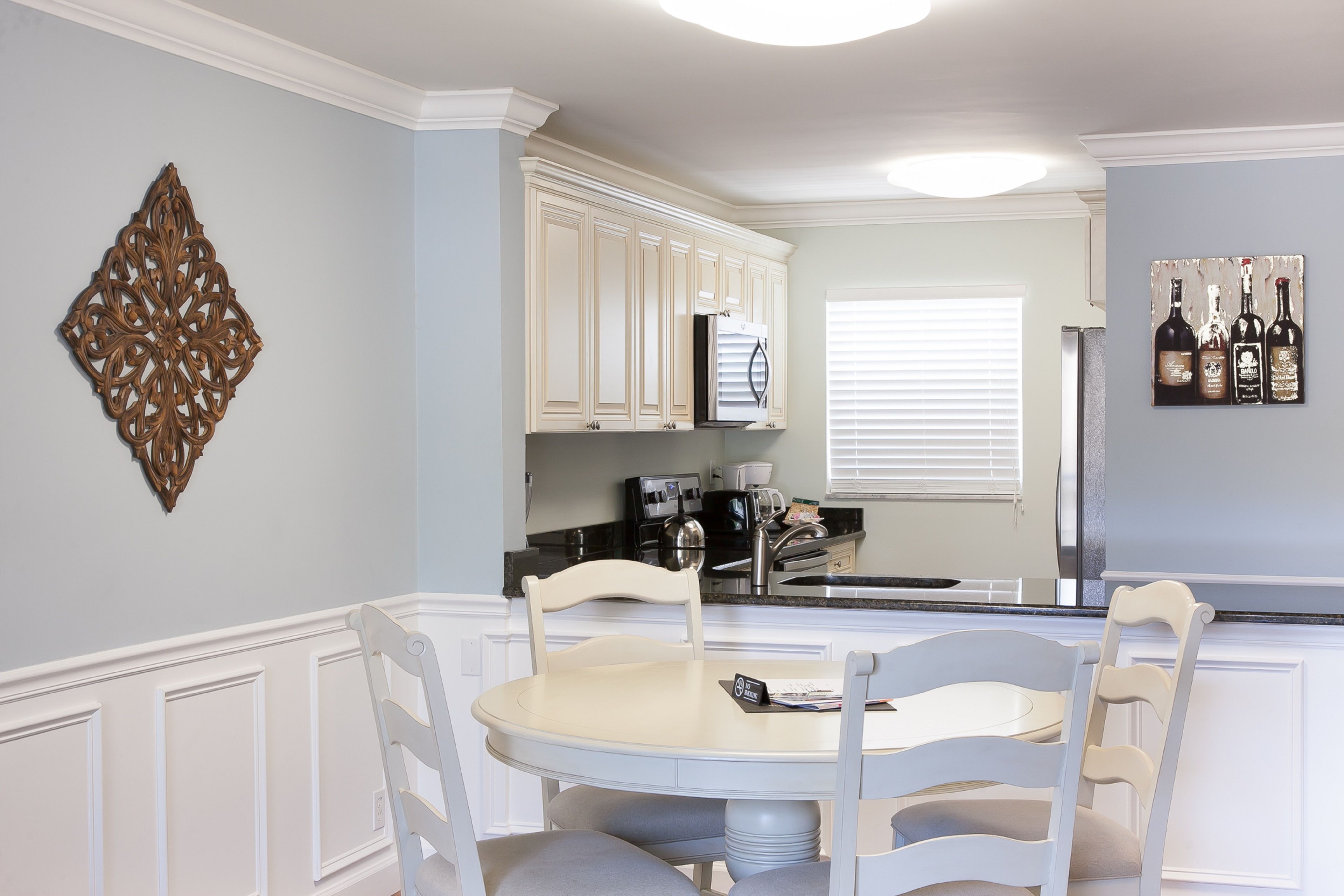 Una cocina con muebles blancos, una mesa de comedor con sillas, un adorno en la pared y una ventana con persianas.