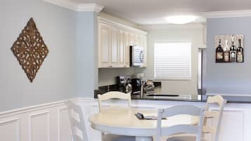 Una cocina con muebles blancos, una mesa de comedor con sillas, un adorno en la pared y una ventana con persianas.