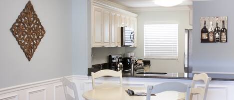 Una cocina con muebles blancos, una mesa de comedor con sillas, un adorno en la pared y una ventana con persianas.