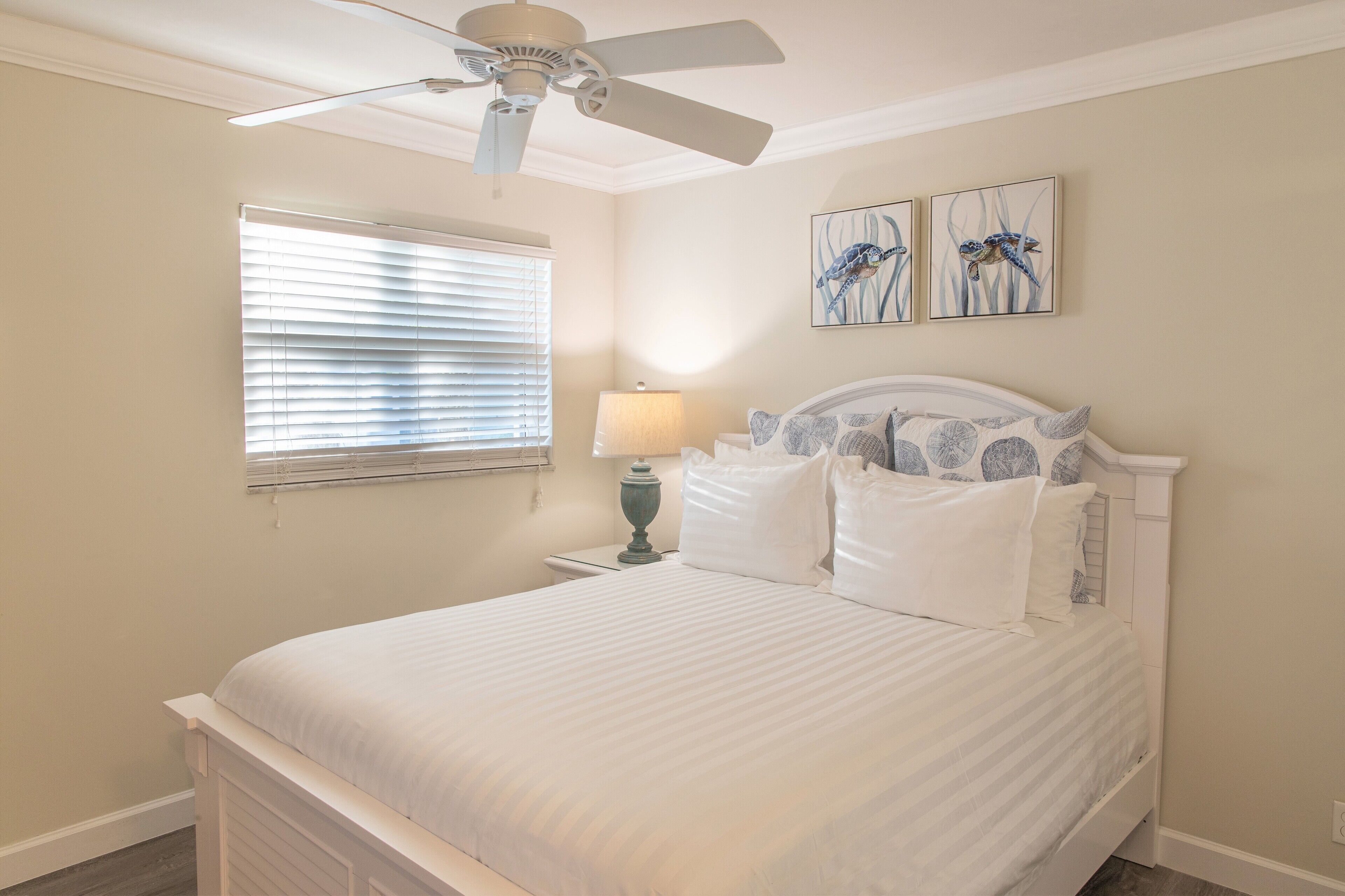 A bedroom with a bed, a ceiling fan, a window with blinds, a bedside table with a lamp, and two wall-mounted paintings.