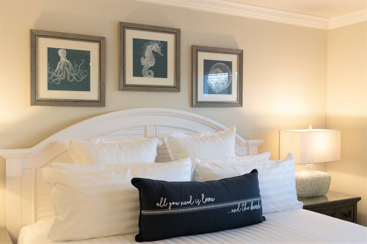 Un lit bien fait, avec du linge de lit blanc et un coussin noir orné d’un motif, une tête de lit en bois et trois œuvres d’art encadrées accrochées au mur.