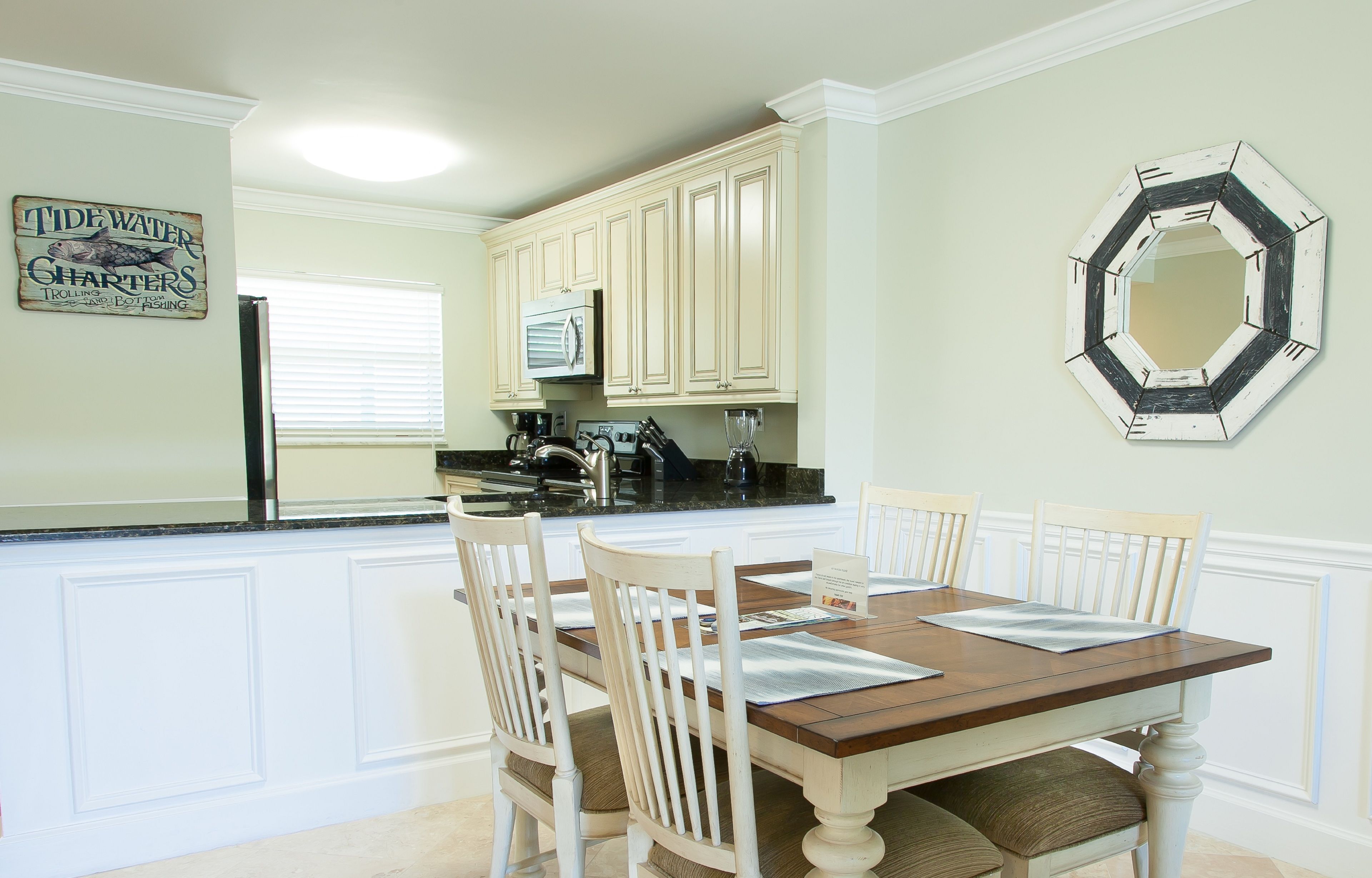 A dining area with a wooden table and white chairs, a kitchen with light-colored cabinets, and a decorative octagonal mirror on the wall.