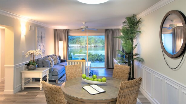 A dining area with a round table, wicker chairs, a bowl of green apples, and a potted plant.