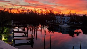 A sunset view from a balcony overlooking a calm body of water with dock structures and buildings in the background. - Dolphin Cove - Adults Only (Freeport)