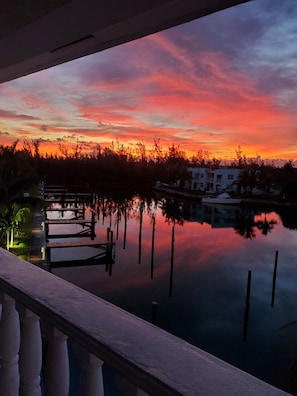 A sunset view from a balcony overlooking a calm body of water with dock structures and buildings in the background. - Dolphin Cove - Adults Only (Freeport)
