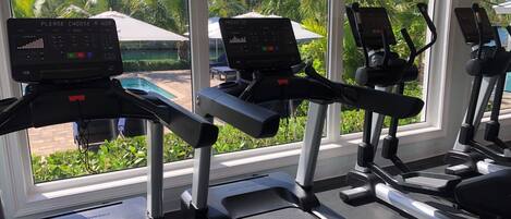Un gimnasio con cintas de correr y equipo de ejercicio, ventanas amplias con vistas a una piscina y palmeras.