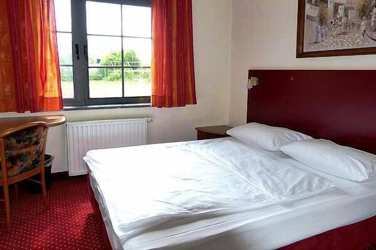 A hotel room with a bed, a chair, a window with curtains, and a painting on the wall.