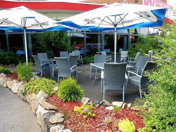 An outdoor seating area with tables and chairs under umbrellas, surrounded by plants and a stone border. - WEST Hotel Radebeul (Radebeul)