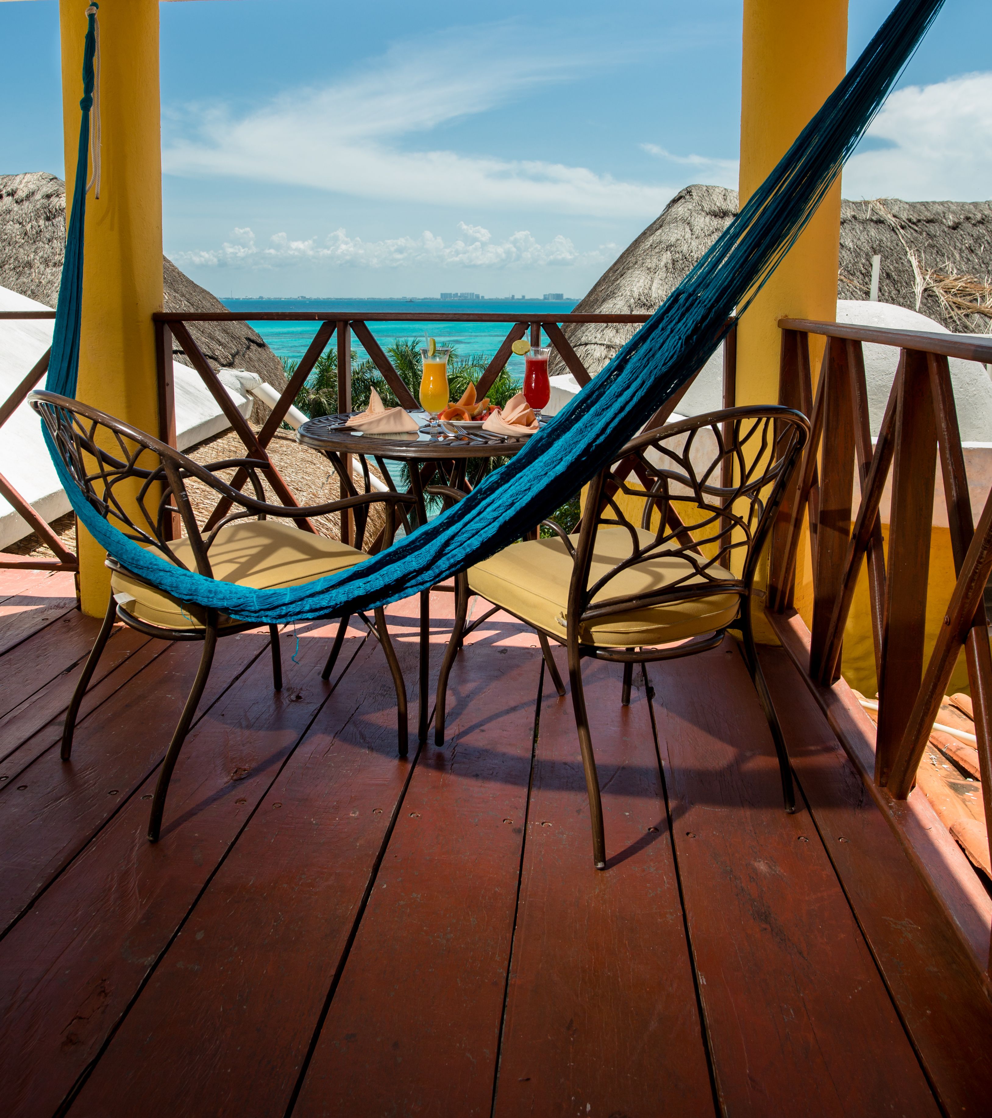 A hammock set up on a wooden deck with a table and chairs, overlooking a beach with a clear view of the ocean.