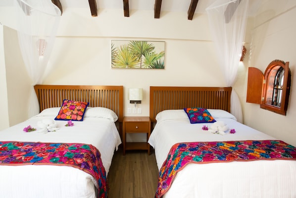 A hotel room with two beds, each with a colorful floral bedspread and matching pillows. There is a nightstand with a lamp between the beds, and a painting of tropical leaves on the wall. - Hotel La Joya (Isla Mujeres)