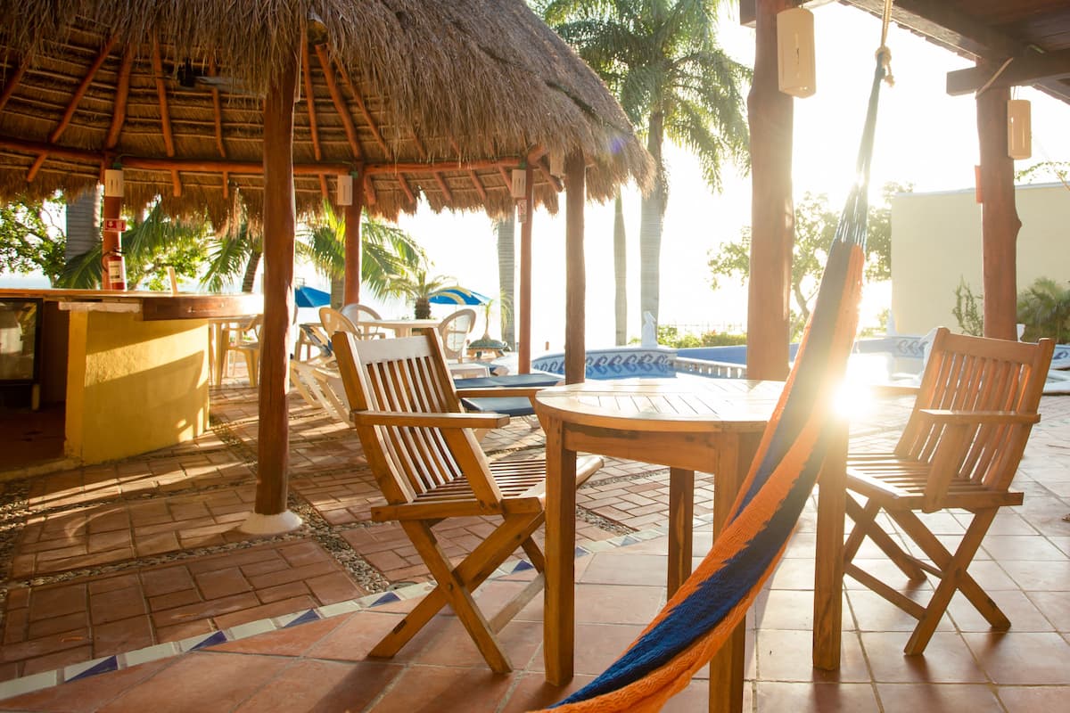 A wooden outdoor seating area with a thatched roof, a pool, and palm trees in the background.