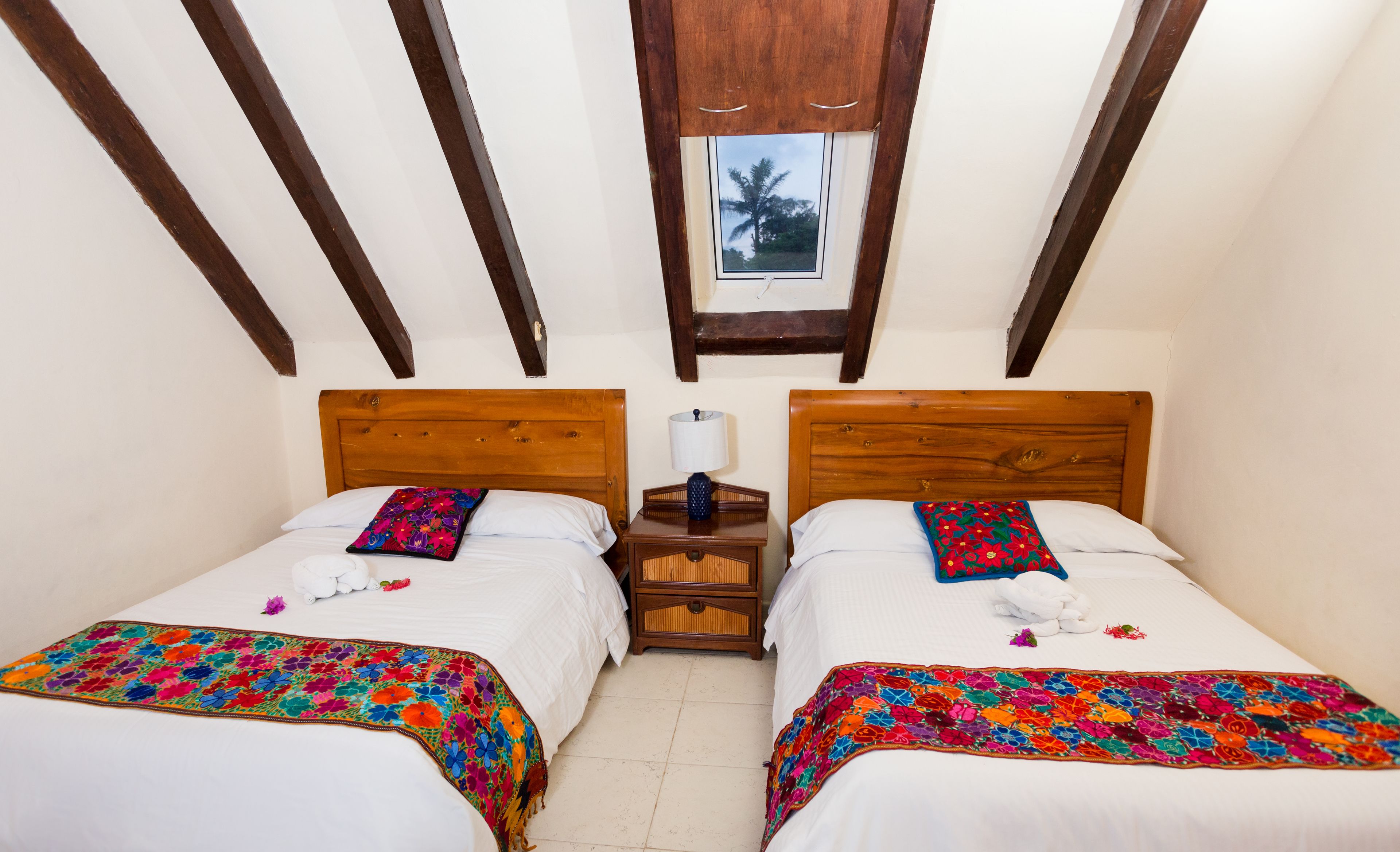 A room with two beds, wooden headboards, a small bedside table with a lamp, and a window with a view of palm trees.