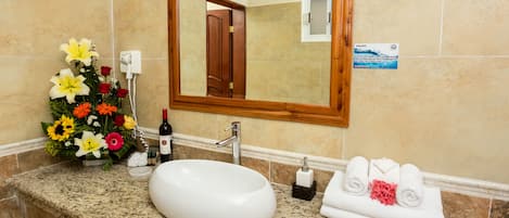 Une salle de bain avec un comptoir en granit, un lavabo blanc, un miroir à cadre en bois et un arrangement floral.