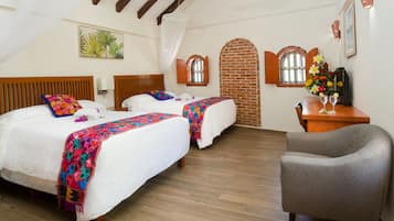 A bedroom with two beds, wooden floors, a brick wall, and a small desk with a vase of flowers.