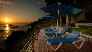 Una terraza de madera con sillones de descanso y sombrillas, con vista a un cuerpo de agua y un atardecer de fondo.