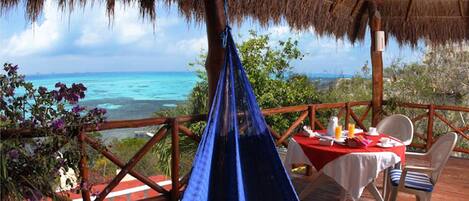 A hammock under a thatched roof, a table with a red tablecloth, and a view of the ocean.