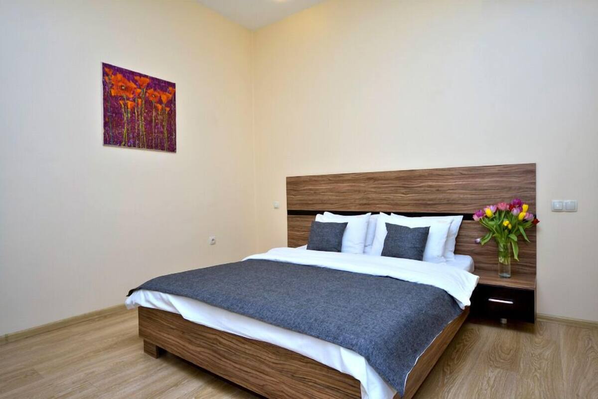 A neatly made bed with a grey blanket, a wooden headboard, and a small bedside table with a vase of flowers.