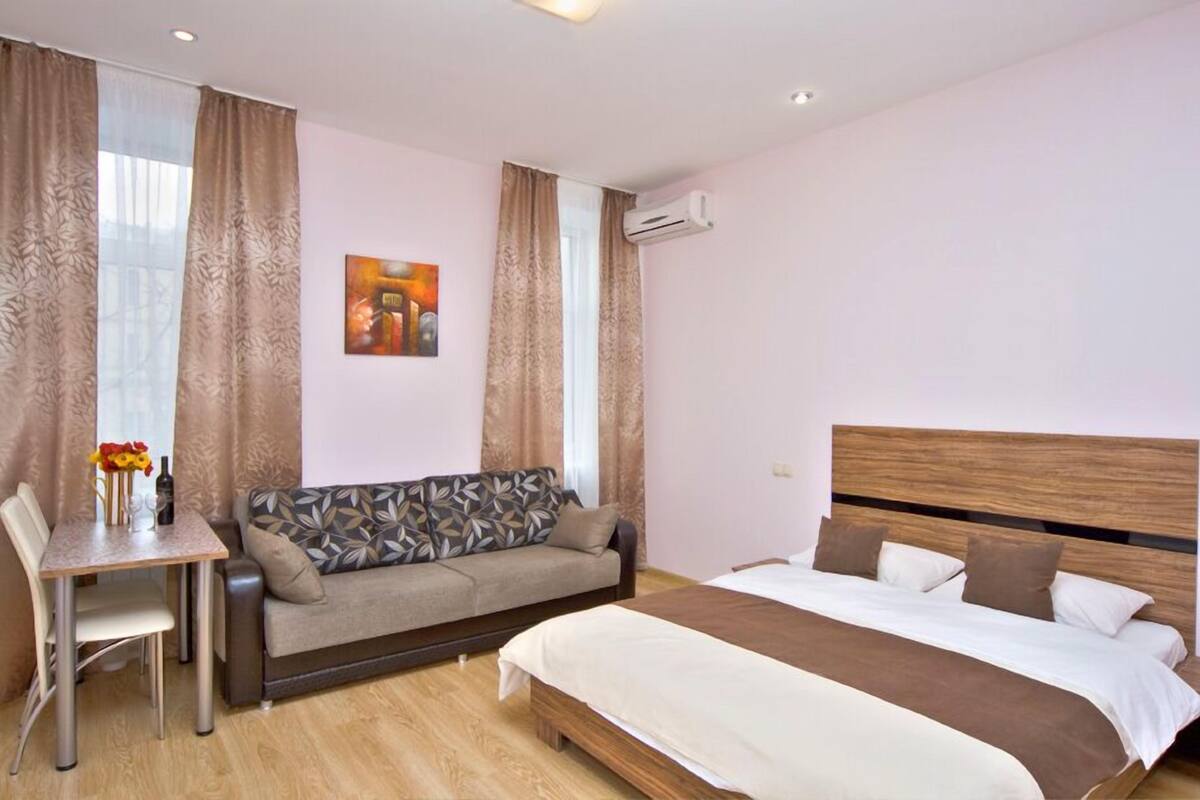A bedroom with a bed, a sofa, a dining table with flowers, and an air conditioning unit.