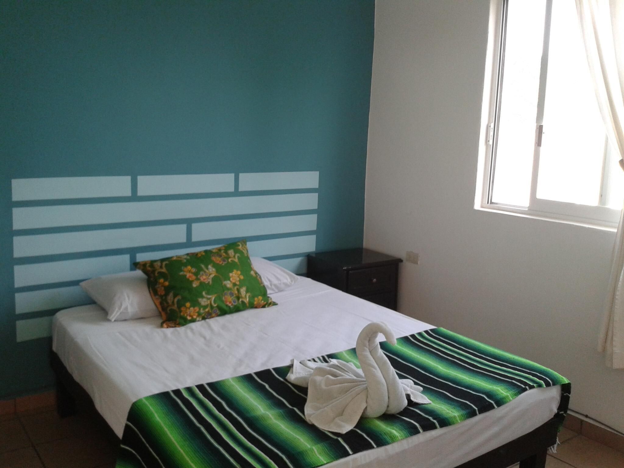 A hotel room with a bed, a green and black striped bedspread, a white swan towel, and a window with curtains.