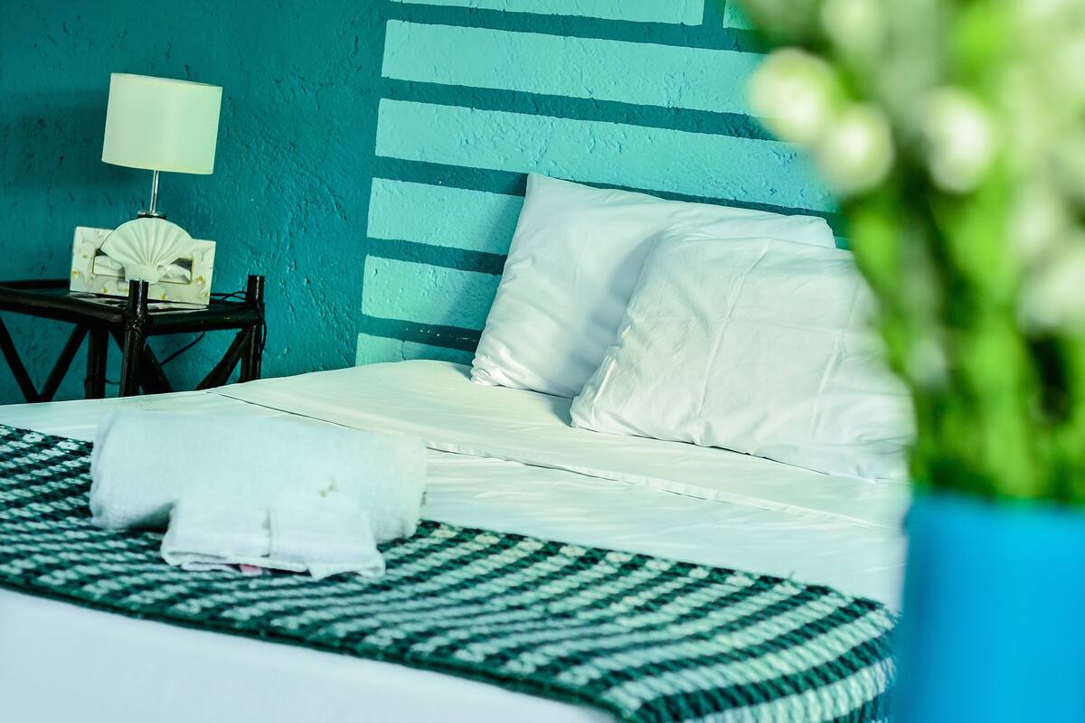 A neatly made bed with white linens, a green and white patterned bedspread, and a white pillow. There is a small bedside table with a lamp and a decorative item. The wall is painted in a teal color with horizontal stripes.