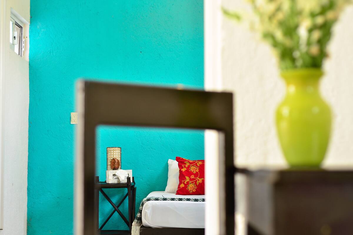 A room with a turquoise wall, a bed with white and red bedding, a small table with a lamp, and a green vase with flowers.