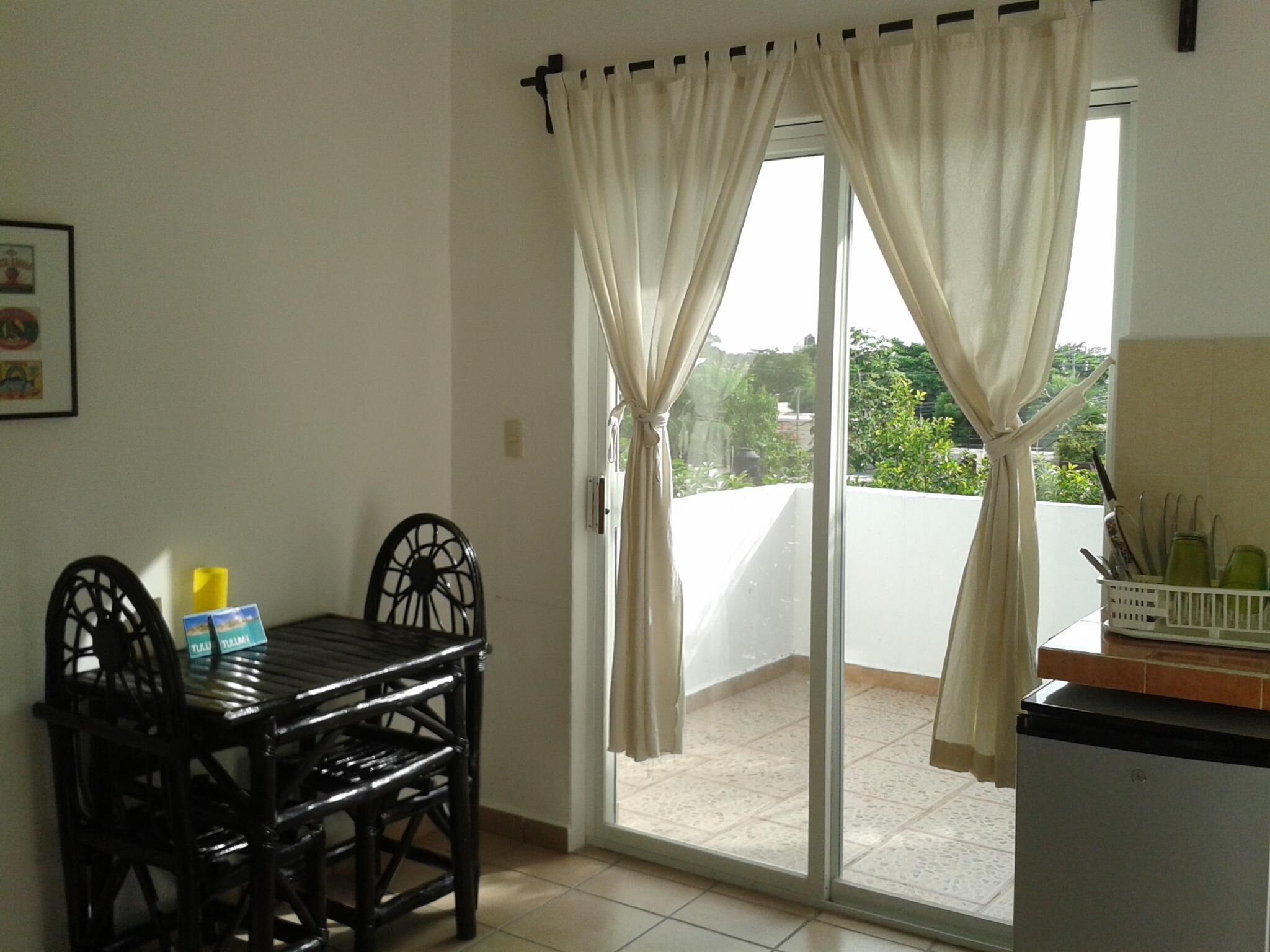 Quarto com mesa de jantar e cadeiras, porta de vidro deslizante com cortinas e uma pequena área de cozinha com utensílios.