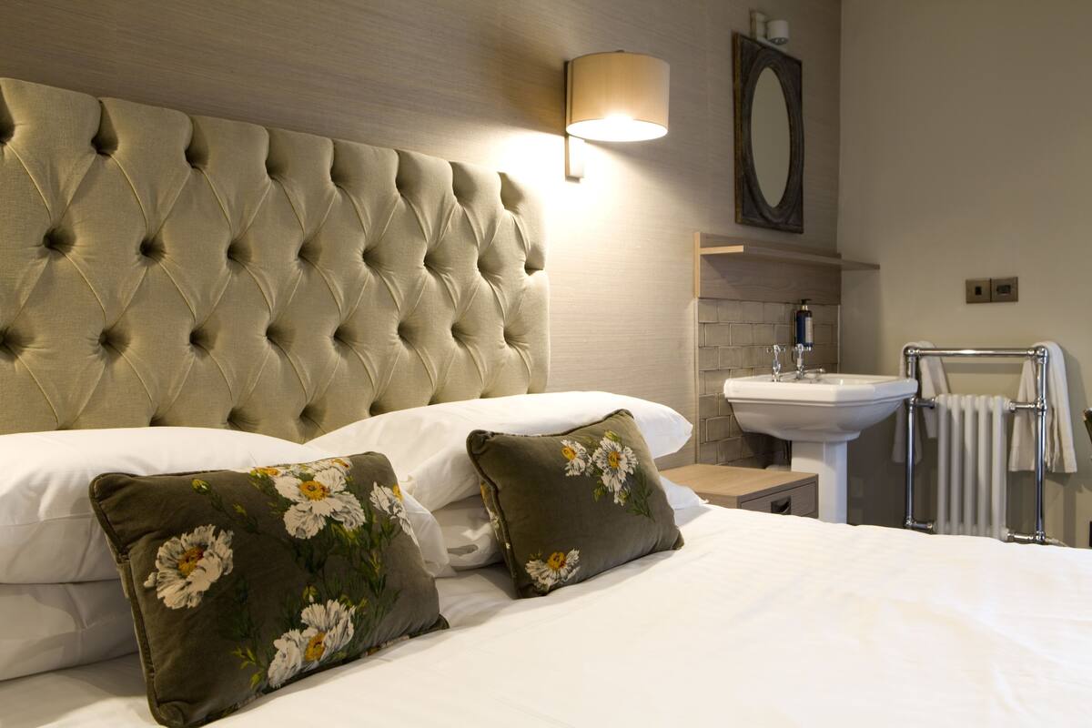 A hotel room with a tufted headboard, a bedside table with a lamp, a sink, and a towel rack.
