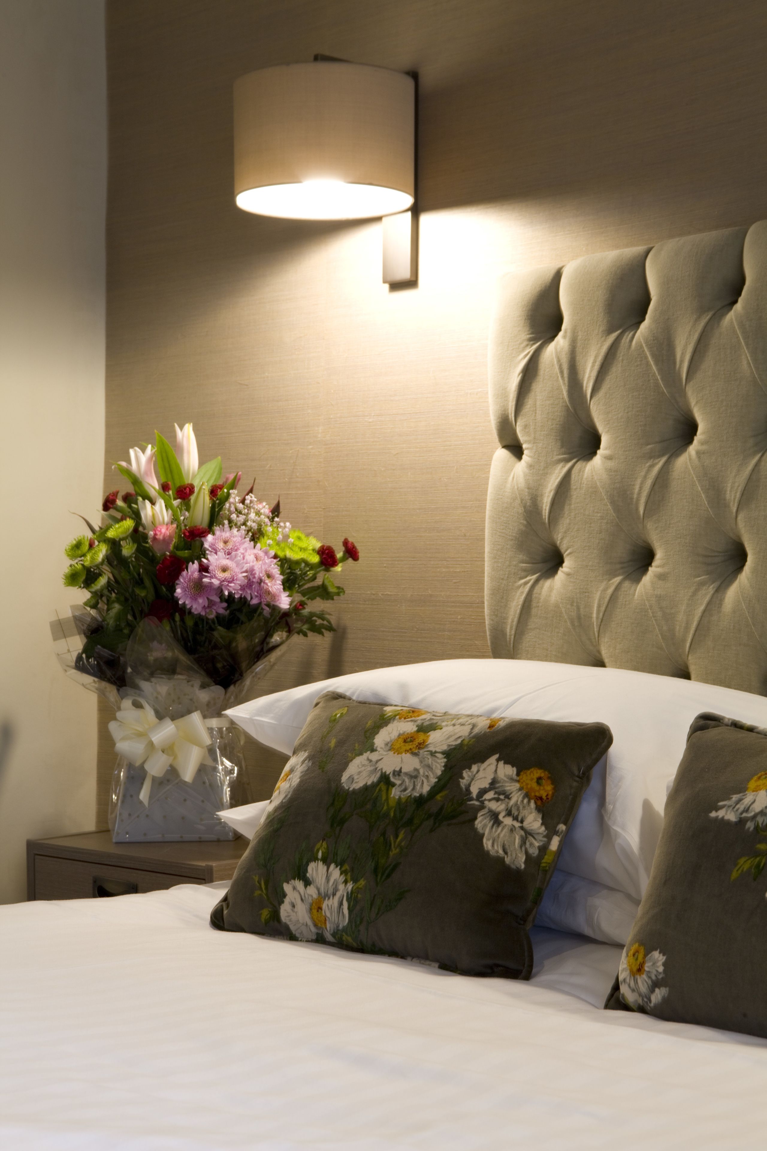 Un lit bien fait, agrémenté d’oreillers à motifs floraux, une lampe de chevet et un arrangement floral posé sur une table de chevet.
