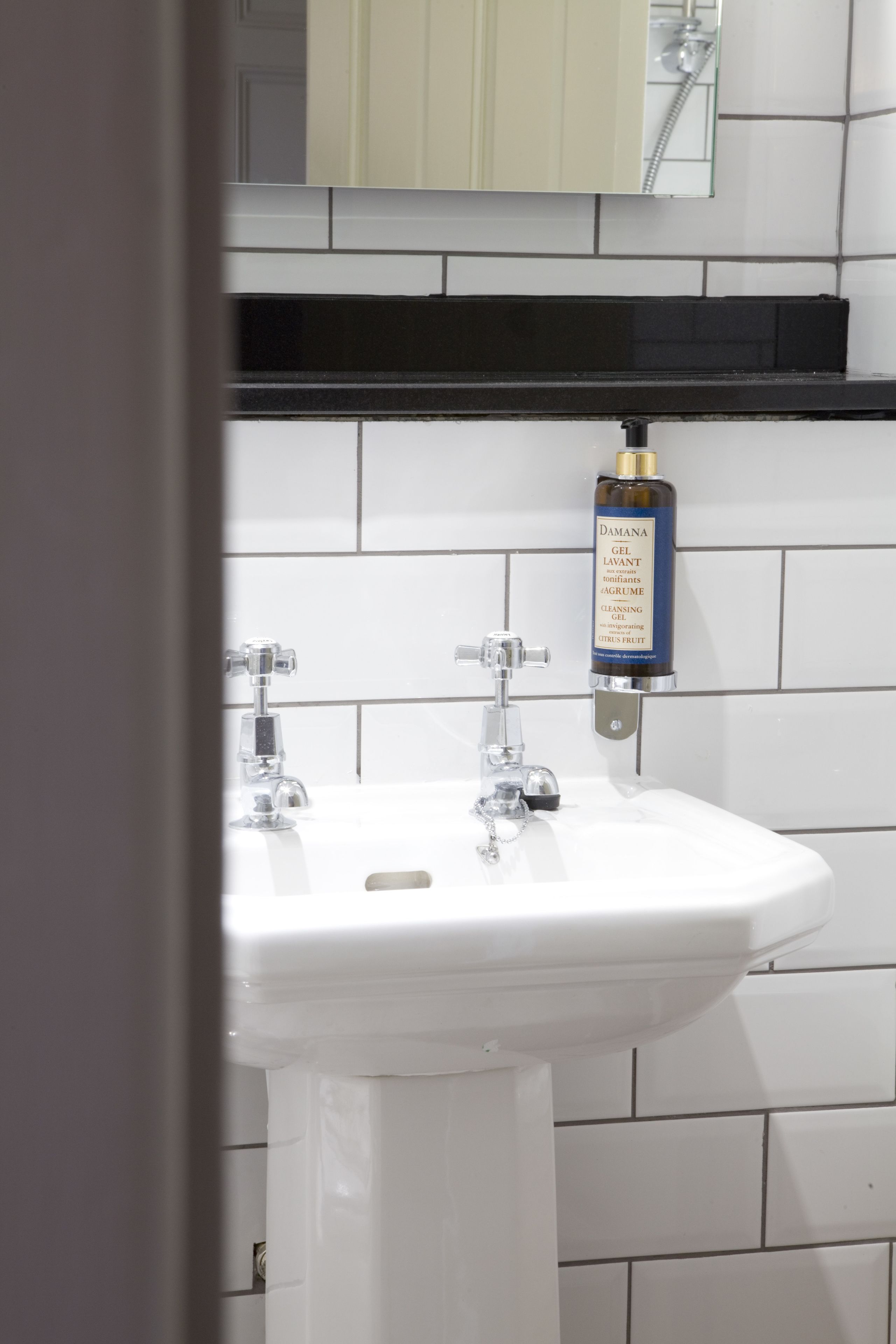A bathroom with a white sink, a mirror above it, and a bottle of DAMANA GEL LAVANT on the wall.