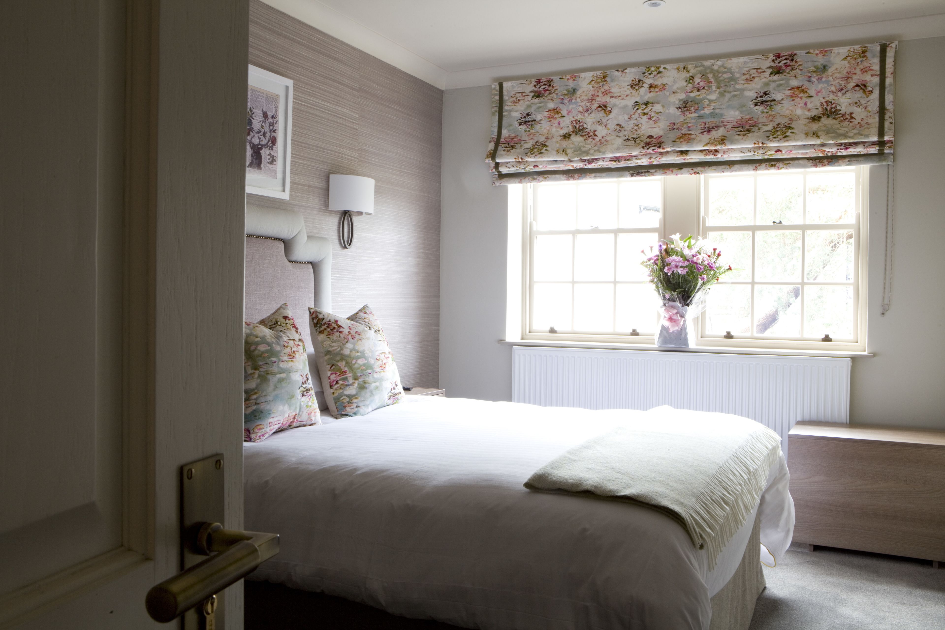 A bedroom with a bed, a bedside table, a lamp, a window with floral curtains, and a radiator.