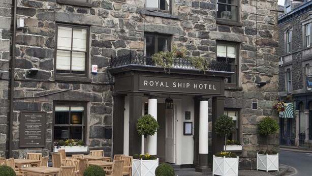 A stone building with the sign "ROYAL SHIP HOTEL" and outdoor seating with white planters.