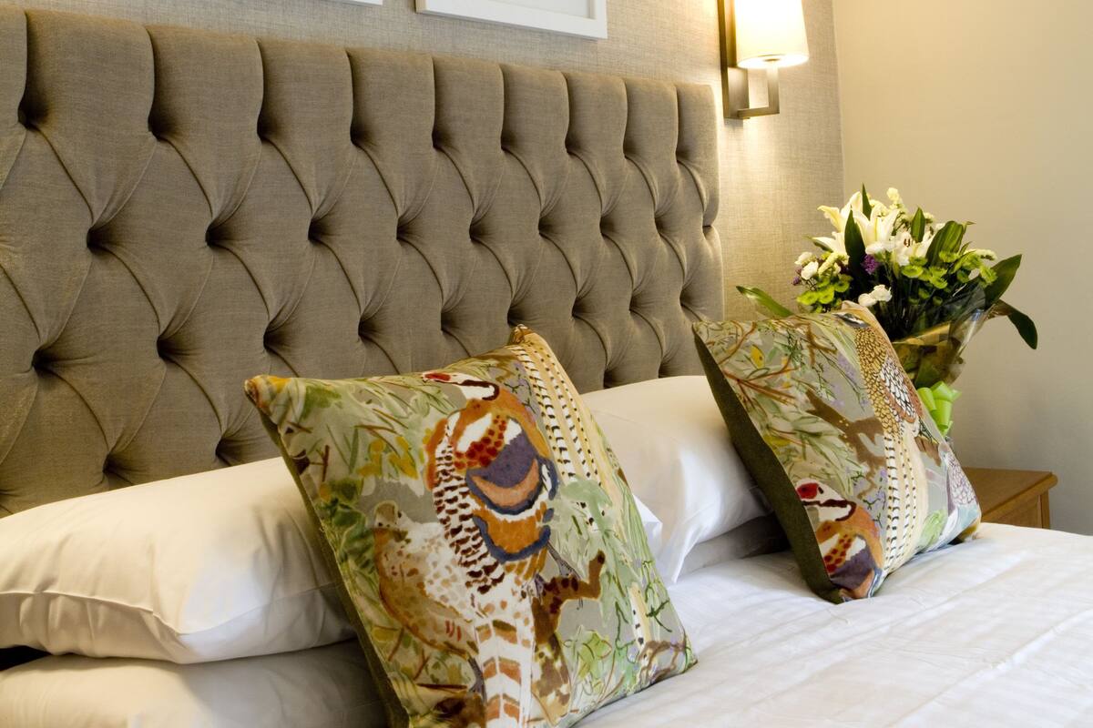 A hotel room with a tufted headboard, two framed pictures on the wall, a bedside lamp, and a floral pillow.