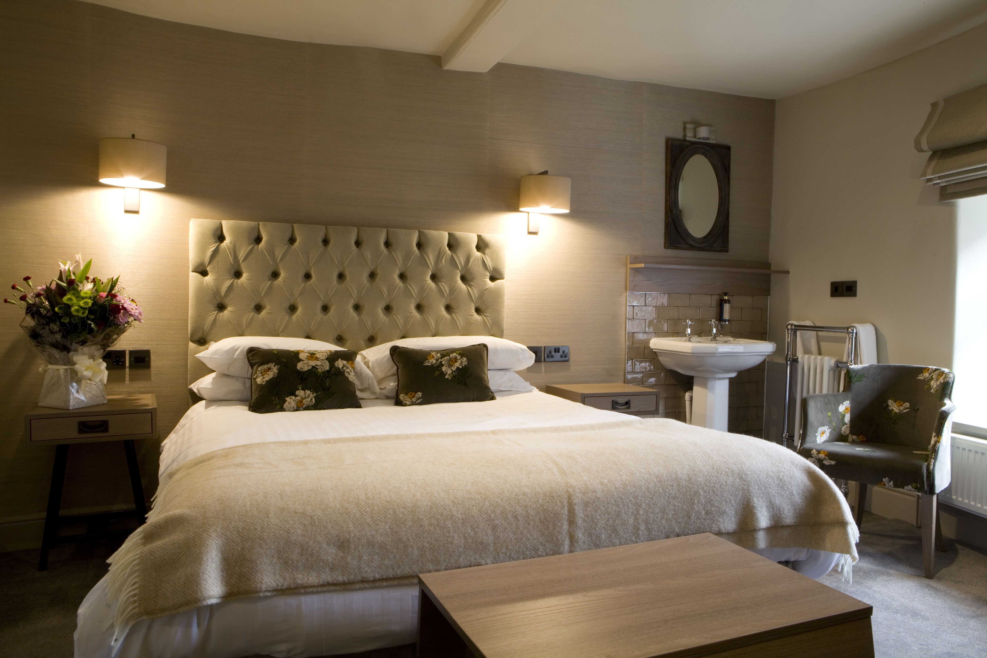 A hotel room with a large bed, a bedside table with a vase of flowers, a chair, a small table, a wall-mounted lamp, a mirror, and a sink.