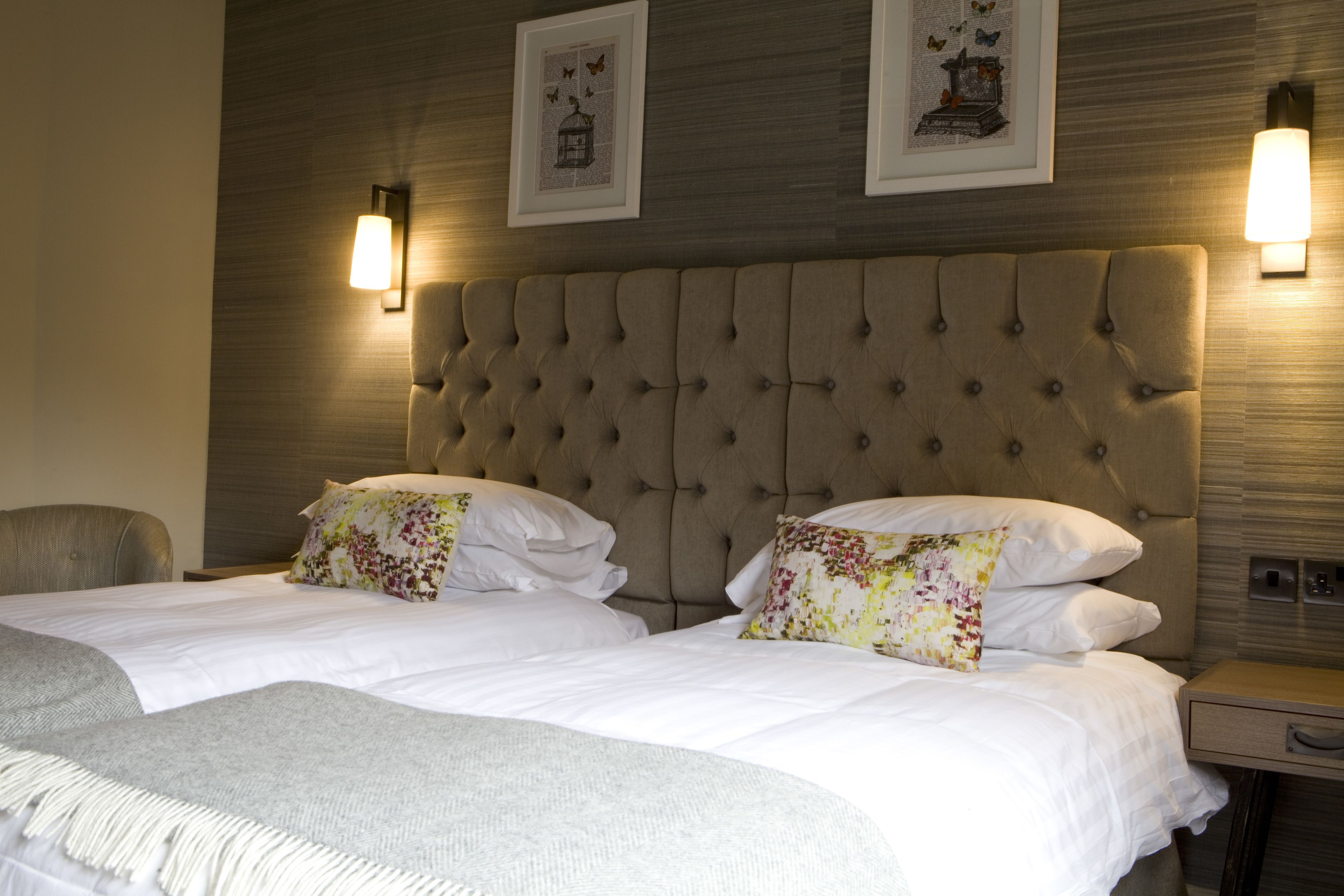 Une chambre d’hôtel dotée d’une grande tête de lit capitonnée, de deux lits avec des oreillers à motifs floraux, d’un plaid gris et de lampes fixées au mur.