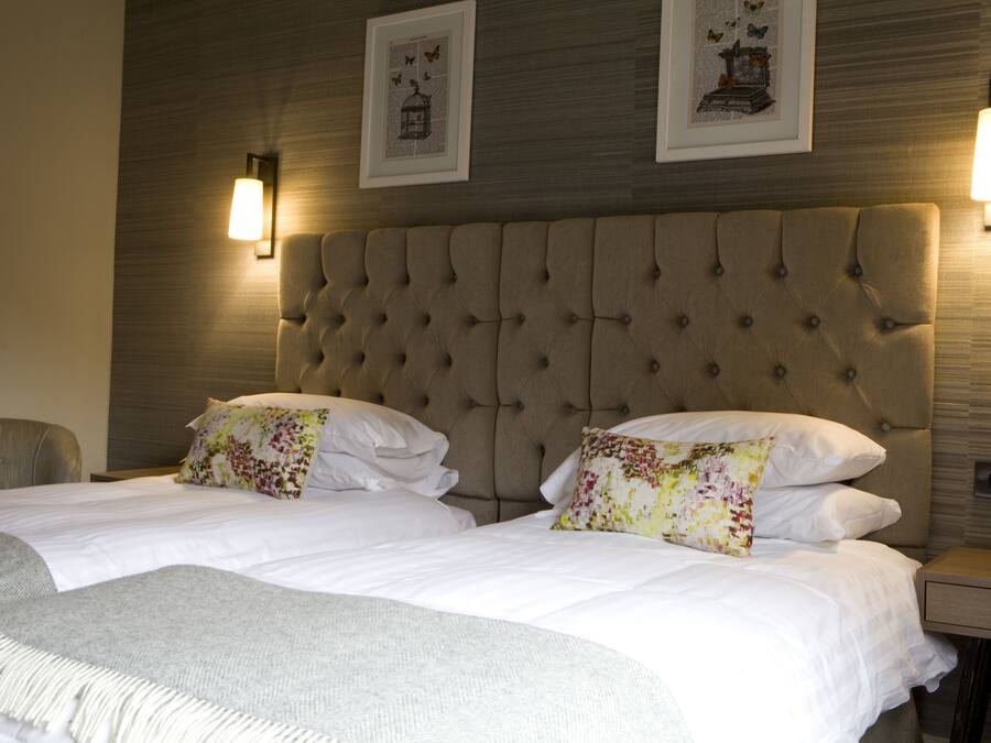 Une chambre d’hôtel dotée d’une grande tête de lit capitonnée, de deux lits avec des oreillers à motifs floraux, d’un plaid gris et de lampes fixées au mur.