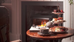 A three-tiered tea service on a wooden table with a chair, a fireplace, and decorative items in the background.