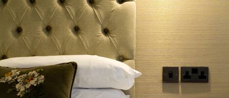A neatly made bed with a tufted headboard, a bedside lamp, and a nightstand with two electrical outlets.
