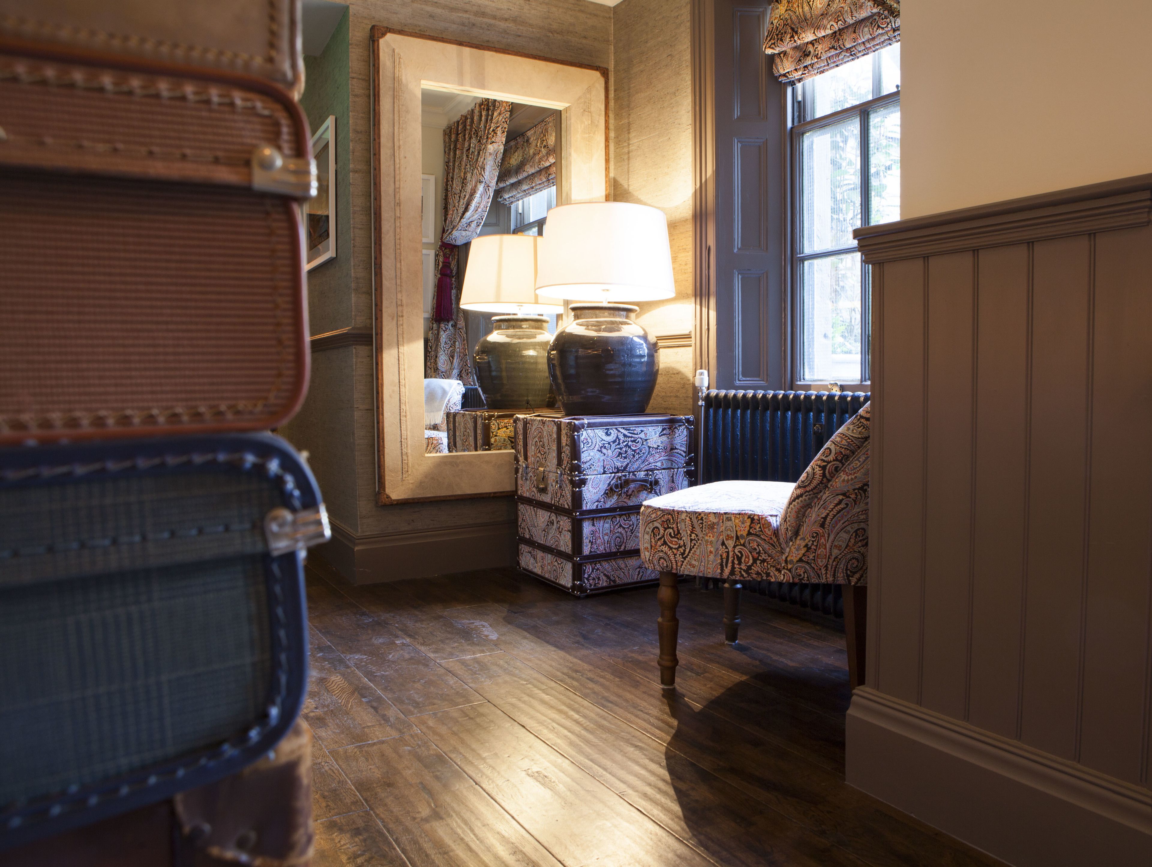 A room with a wooden floor, a chair, a lamp, a large vase, and a mirror reflecting a window with curtains.