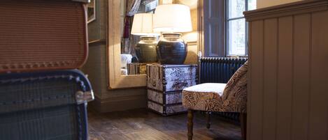 A room with a wooden floor, a chair, a lamp, a large vase, and a mirror reflecting a window with curtains.