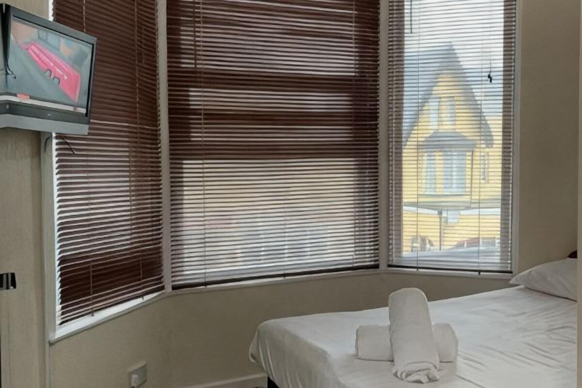 Una habitación de hotel con una cama, un televisor montado en la pared y ventanas grandes con persianas.
