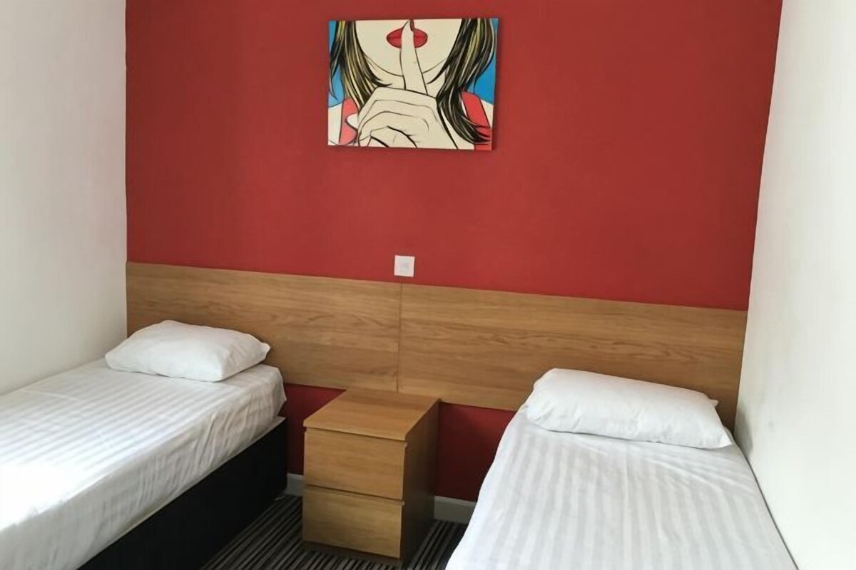 Una habitación con dos camas individuales, un cabecero de madera, una mesita de noche de madera y una pared roja con un cuadro.