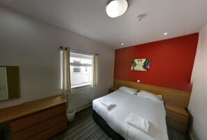 Een hotelkamer met een bed, een dressoir, een raam met gordijnen, een rode accentmuur en een plafondlamp.