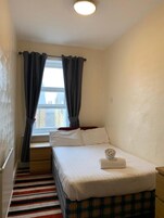Een kleine, netjes ingerichte slaapkamer met een enkel bed, een raam met gordijnen, een gestreept tapijt en een effen muur.