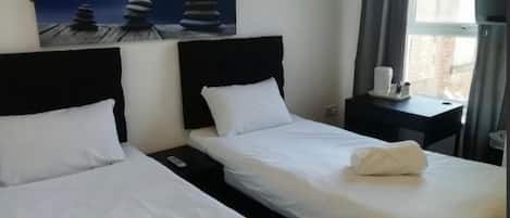A hotel room with two beds, a painting of stacked stones, a nightstand with a remote control, and a window with curtains.