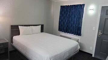 A hotel room with a bed, a bedside table with a phone, a window with blue curtains, and a door.